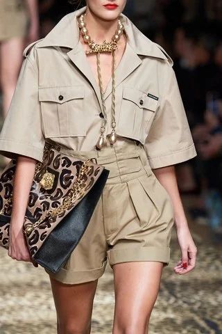 a fashion model walks the runway showcasing a stylish summer outfit. she is wearing a beige khakicolored shortsleeved shirt and highwaisted pleated shorts.  a large, statement necklace with pearls and a gold pendant is a key accessory. she carries a black and leopardprint clutch. the overall style is sophisticated and summery. the background is blurred, focusing attention on the model and her outfit.