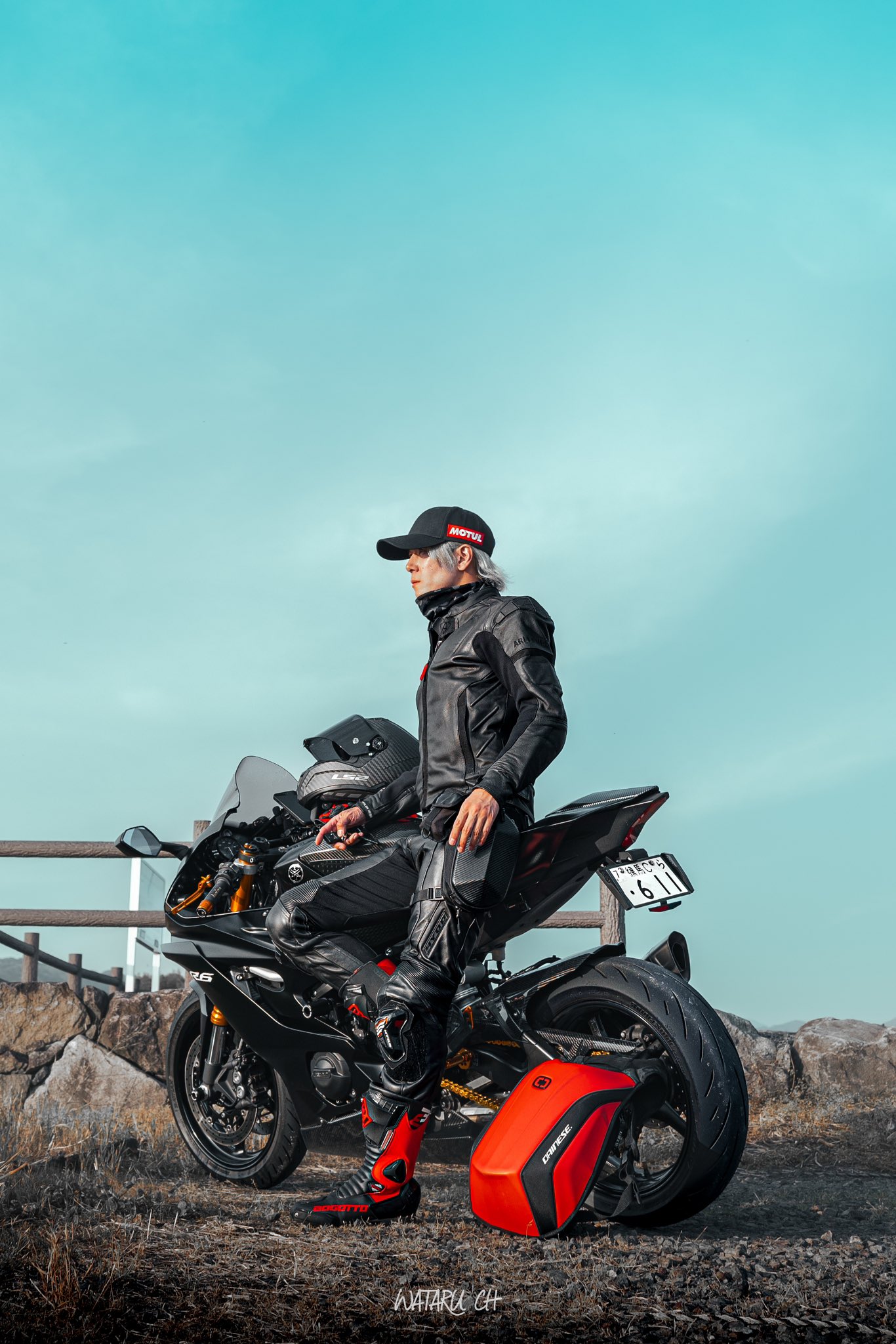 a biker in a black leather jacket and a black helmet sits on a black yamaha r1 motorcycle, looking out at the blue sky. the motorcycle has a red tank bag and a red tail bag. the biker is wearing black motorcycle boots.