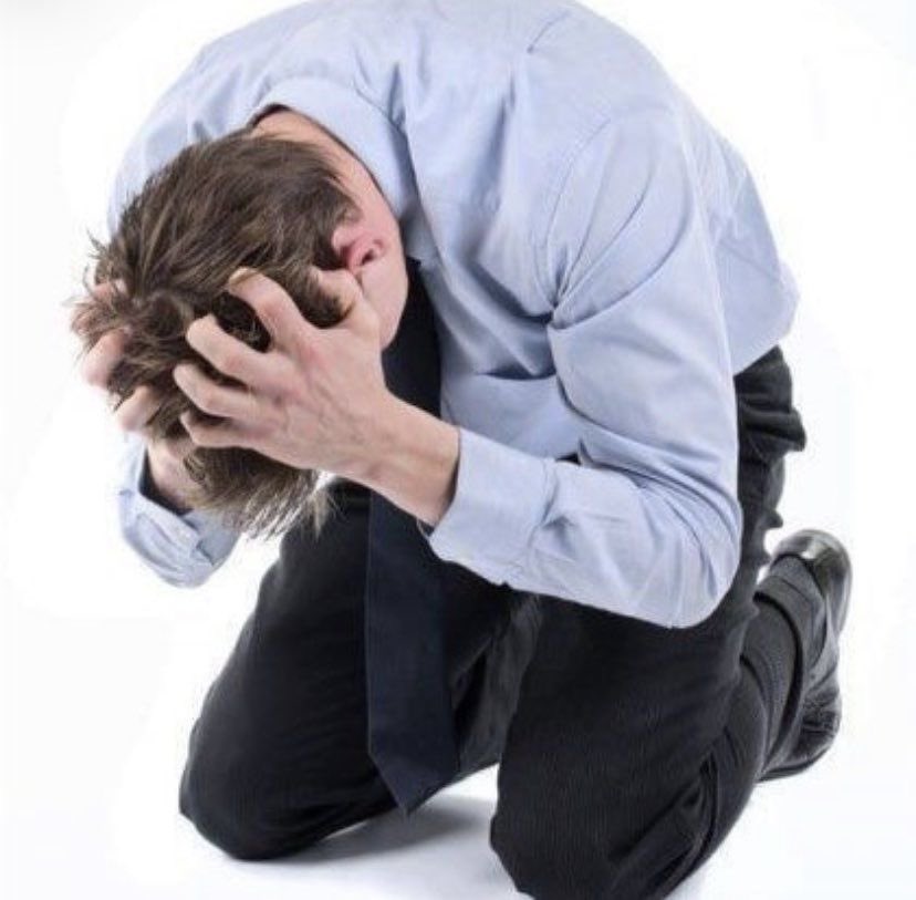 a distressed businessman is kneeling with his head in his hands. he appears upset, overwhelmed, and possibly depressed. he is wearing a light blue shirt, a dark tie, and dark pants. the image evokes feelings of despair and anxiety.