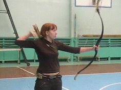 a woman, with red hair pulled back, is captured midarchery pose in a brightly lit gymnasium. she is positioned slightly to the left of center, her body angled as she draws back a recurve bow with her right hand. her focused gaze follows the trajectory of the arrow, already absent from the picture. dressed in black athletic wear, she stands out against the muted green walls and blue flooring, characteristic of an indoor sports facility. the perspective provides a clear view of her form and the intensity of her concentration.