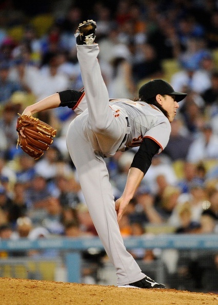 a baseball pitcher in a white uniform with orange and black details is captured midpitch. his left leg is raised high in the air, showcasing the athleticism and flexibility required of the position. he's seen throwing with his right hand, and his glove is still raised from catching the ball in his left. the blurred background suggests a stadium filled with spectators, indicating a live game setting. the image exudes action and energy, highlighting a pivotal moment in the game.