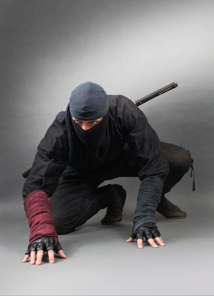 a ninja, dressed in a black ninja suit and a black mask, is crouched on the ground,  his right hand is stretched out with his fingers extended, while his left hand is holding a sword. his face is obscured by the mask and he is looking at the camera. his pose suggests that he is ready to strike.  the image evokes a sense of danger and mystery, as if the ninja is about to disappear into the shadows.
