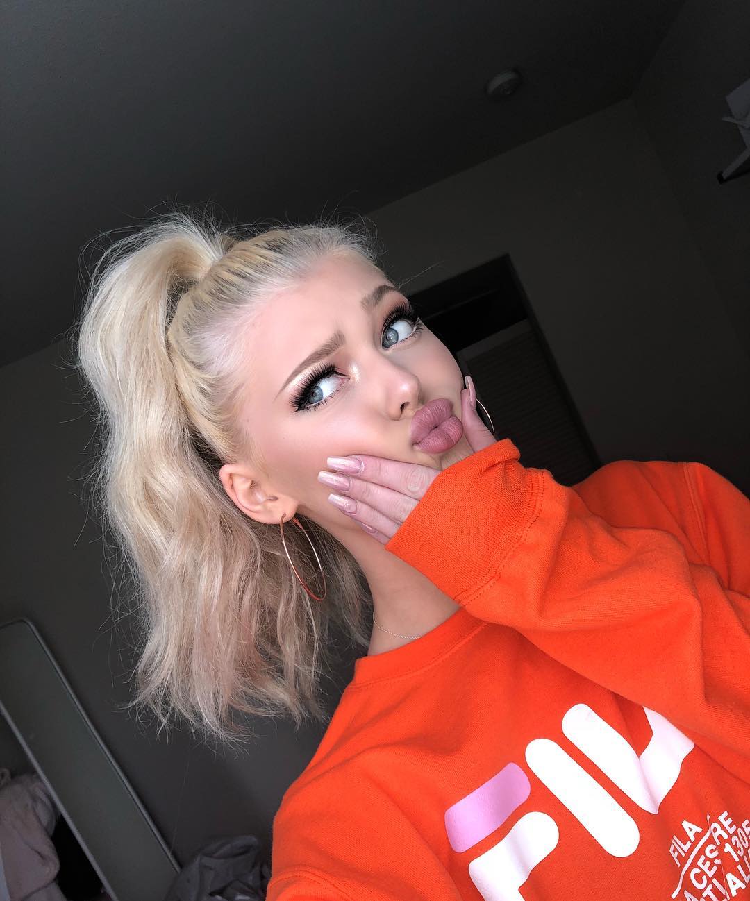 a young woman with blonde hair in a high ponytail is pouting at the camera. she's wearing an orange fila sweatshirt and large hoop earrings. her hand is on her face. the background appears to be a bedroom.