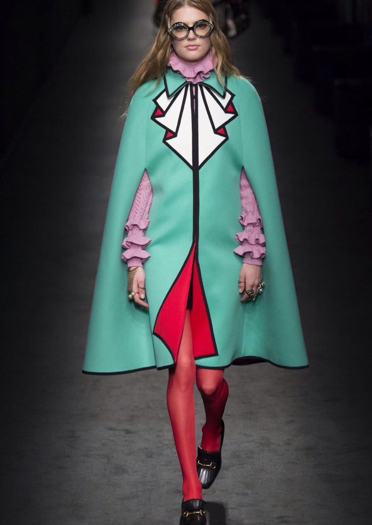 a female fashion model wearing a green cape with a red and black design, a pink bow collar and red tights. the model is on a runway, walking towards the camera. the model is wearing black shoes.  the model is posing, with her hair pulled back and wearing glasses.  the model is surrounded by a dark background. the model is wearing a bright and bold look.