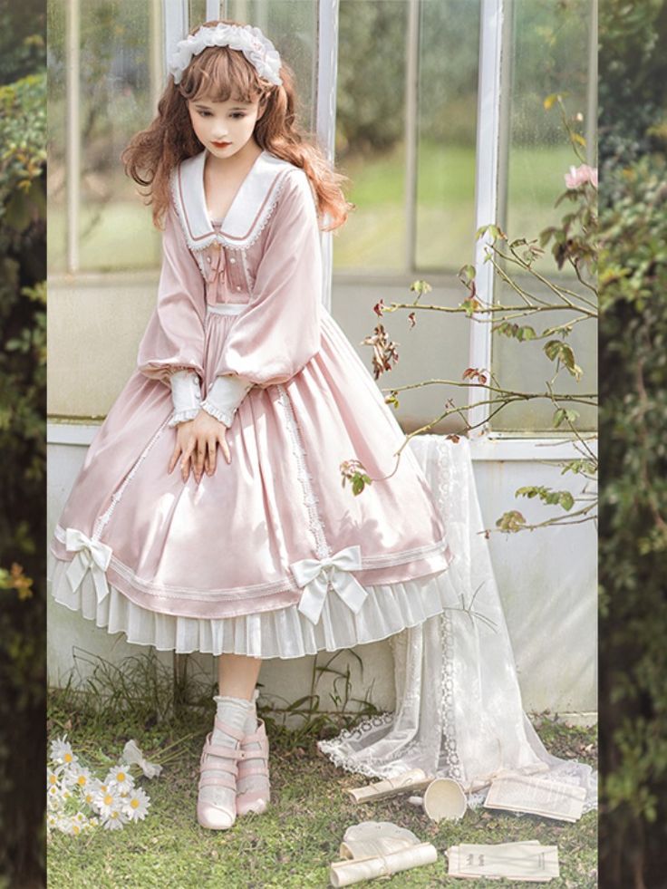a young girl is wearing a pink and white lolita fashion dress and standing in a vintage studio. the dress has long sleeves, a square neckline, and a full skirt. she is also wearing a white headband with a bow, white socks, and pink shoes. she has long brown hair and is looking down at the ground. there are some white flowers on the floor next to her.