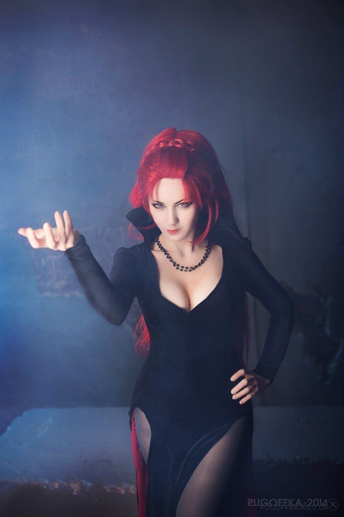 a woman dressed in a cosplay outfit stands in a dimly lit, smoky setting. she has vibrant red hair styled with a braid at the top, and wears a black dress with a deep vneck, exposing cleavage, and a thighhigh slit. a black beaded necklace adorns her neck. her makeup is striking, and she poses with one hand open as if casting a spell, while the other rests on her hip. the background is obscured by smoke and darkness, creating a fantasy or gothic ambiance.