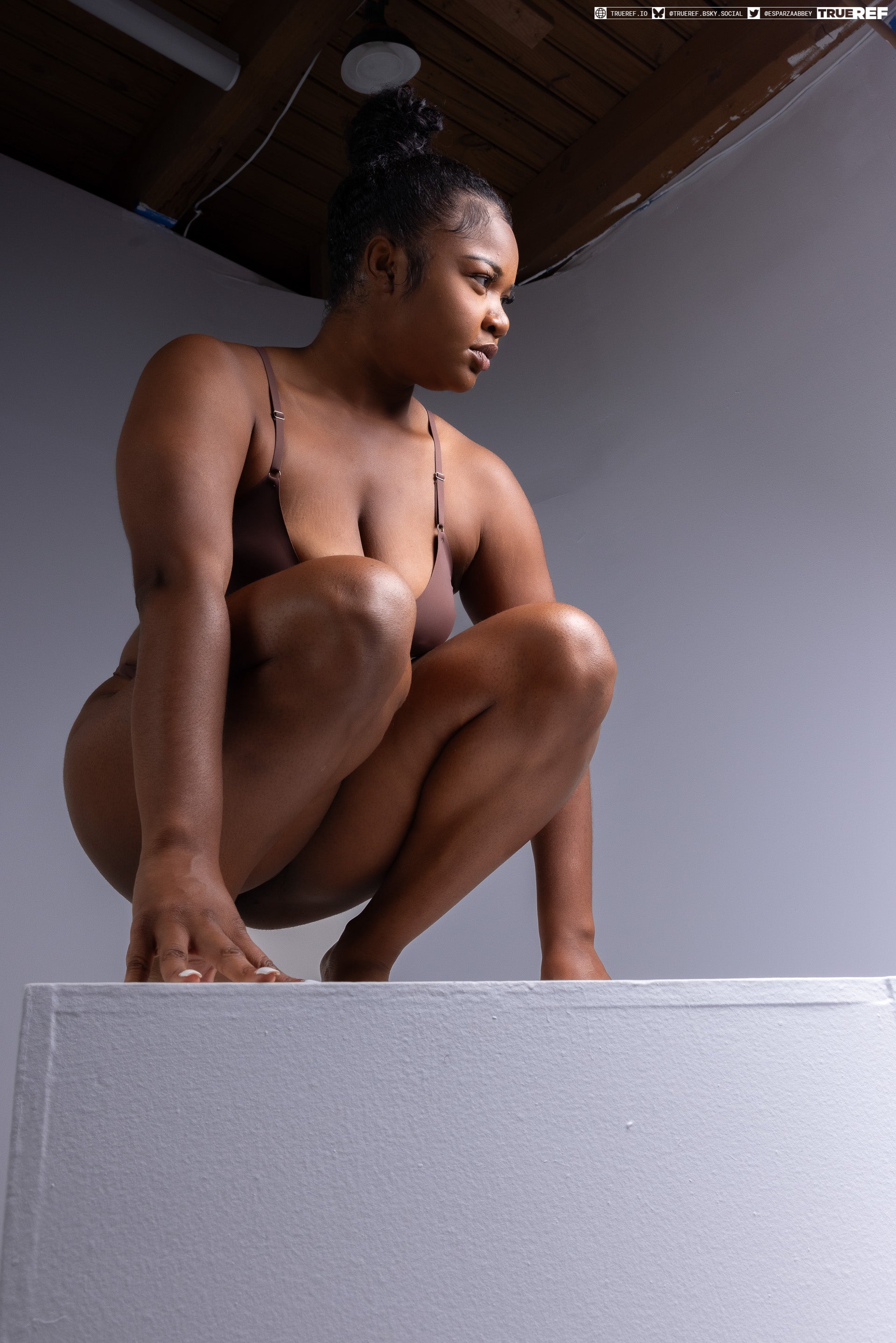 a woman with dark hair in a bun is crouching on a white platform. she is wearing underwear and looking away from the camera. the background is a grey wall and a dark ceiling.