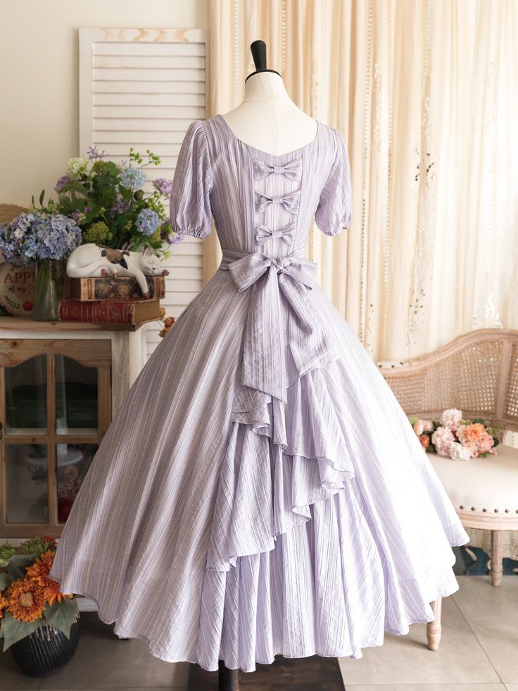 a flowy, purple and white striped dress with a square neckline and puff sleeves. the back of the dress has a large bow and a tiered skirt with ruffles. the dress is displayed on a mannequin in a room with natural light.