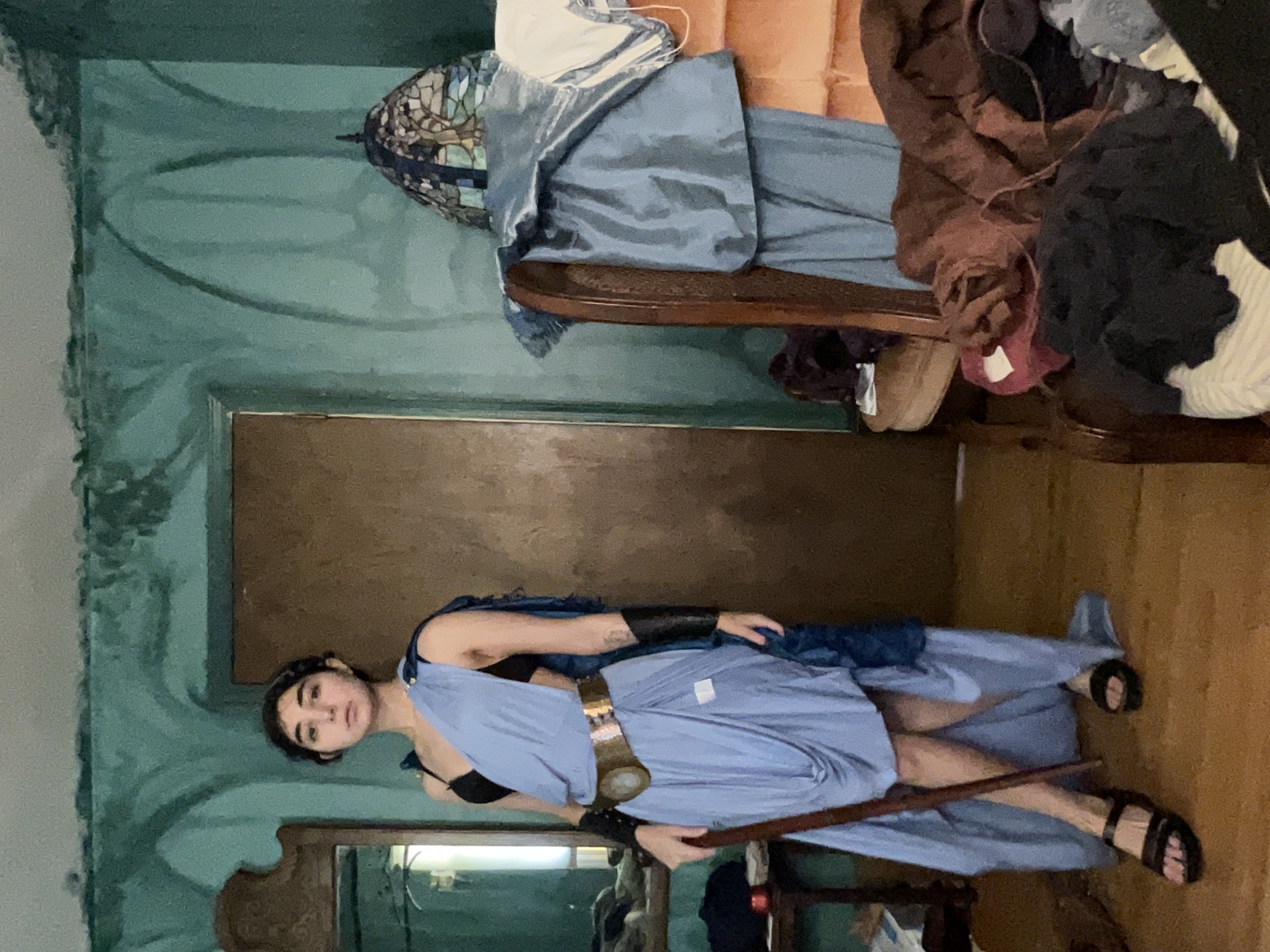 a young woman in a blue grecianstyle cosplay dress stands in a messy room.  she holds a wooden staff and is wearing sandals. the room has teal walls, antique furniture including a chair piled with clothing, and a vintagestyle stainedglass lamp. the overall aesthetic is a blend of fantasy and slightly disheveled reality.