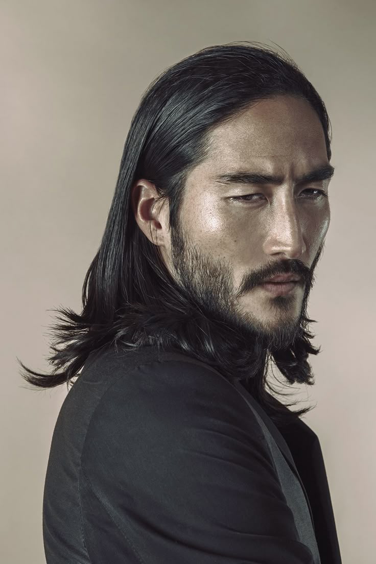 a portrait of an asian man with long, dark hair and a beard and mustache, wearing a black shirt. he has an intense, serious expression and is looking slightly to the side.