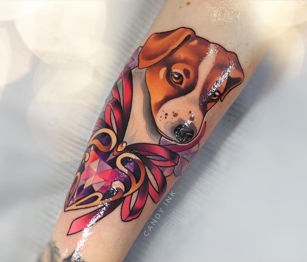 a neo traditional tattoo of a jack russell terrier's face. the dog is posed slightly in profile and wears a red ribbon tied around its neck. the ribbon is attached to a heart shaped pendant with a pink and purple gemstone.  this tattoo appears on someone's inner forearm.