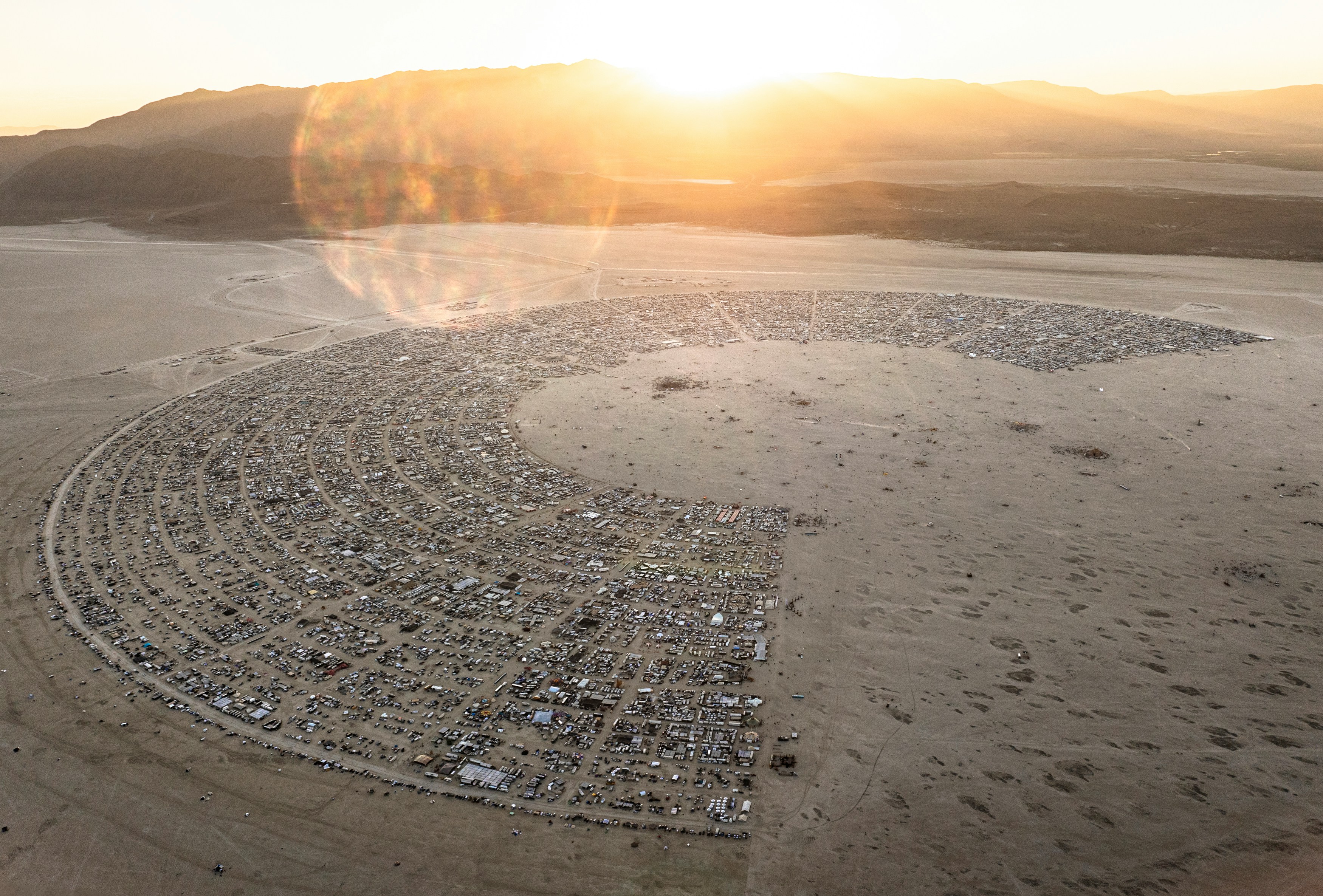an aerial view of black rock city, the temporary city erected annually for the burning man festival. the city is laid out in a circular pattern, with a large open space in the center. thousands of tents and vehicles are visible, indicating the large number of people attending the festival. the sun is setting in the background, casting a warm golden light over the scene. the surrounding landscape is barren desert, with mountains visible in the distance.