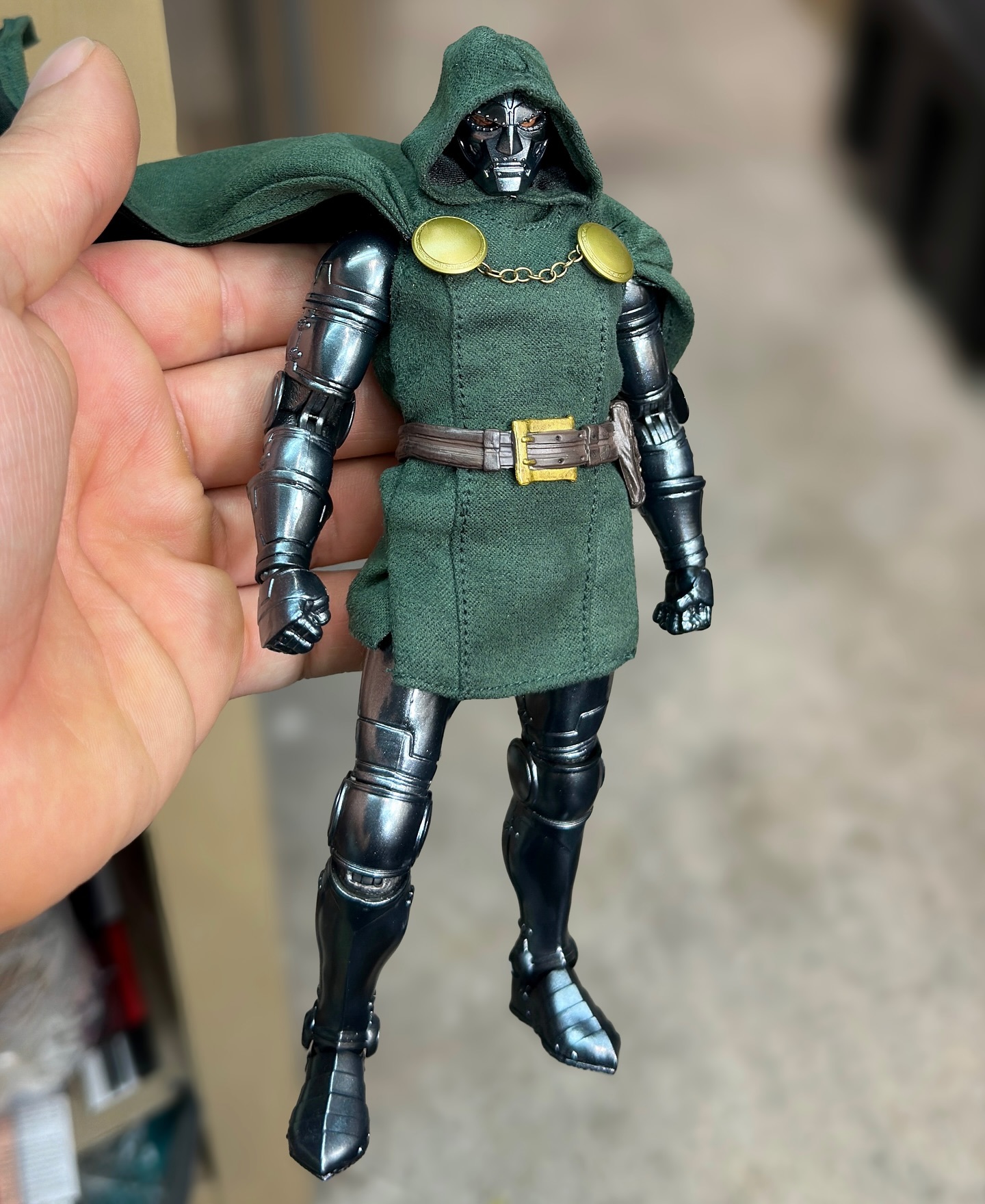 the image shows a doctor doom action figure being held in a hand. the figure has a green cloak, metal armor, and a menacing expression. it is likely a collectible item or toy.