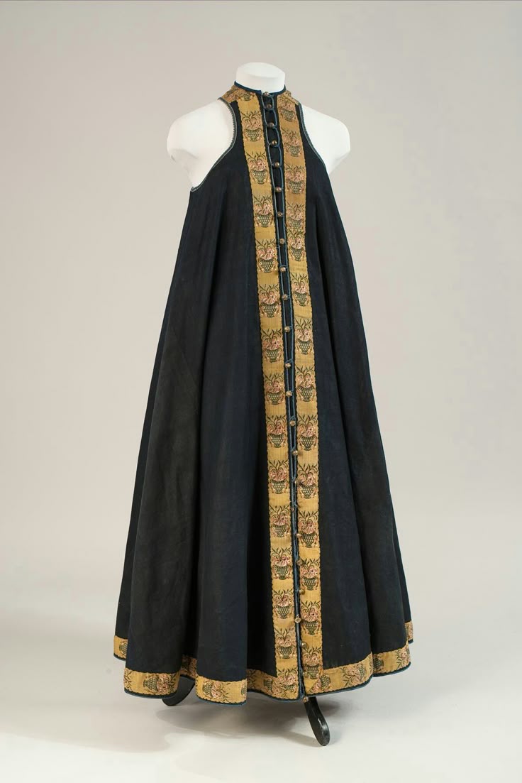 a fulllength studio shot shows a dark, sleeveless dress with floral pattern trim on a mannequin set against a neutral background. the dress has a high neck and straight cut down the front with the floral trim running along the front from neck to hem and also on the hem of the dress. the trim has a brown background with small floral patterns in gold and pink. the dress is buttoned down the front. the mannequin is white and featureless.