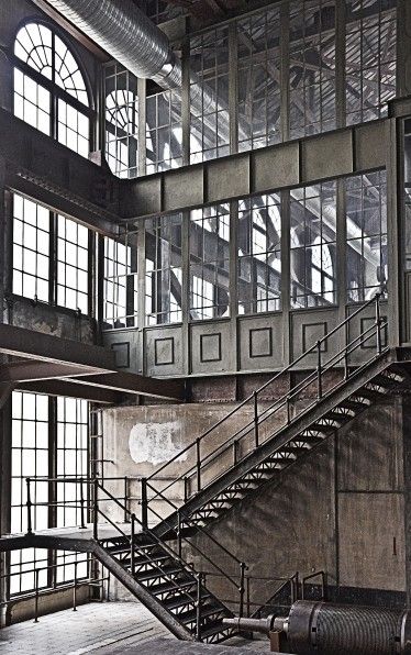 the image shows the interior of a large industrial building, possibly a factory or power plant.  the scene is dominated by a long metal staircase that descends diagonally across the frame. the building features expansive windows allowing significant natural light to enter, contrasting with the aged and somewhat weathered look of the interior. the walls are made of concrete and brick, and various structural elements like pipes and metal beams are visible.  a large piece of industrial machinery is partially visible in the bottom right corner. the overall atmosphere is one of industrial decay and beauty, with a strong emphasis on the interplay of light and shadow.  the image hints at the building's past significance and industrial heritage.