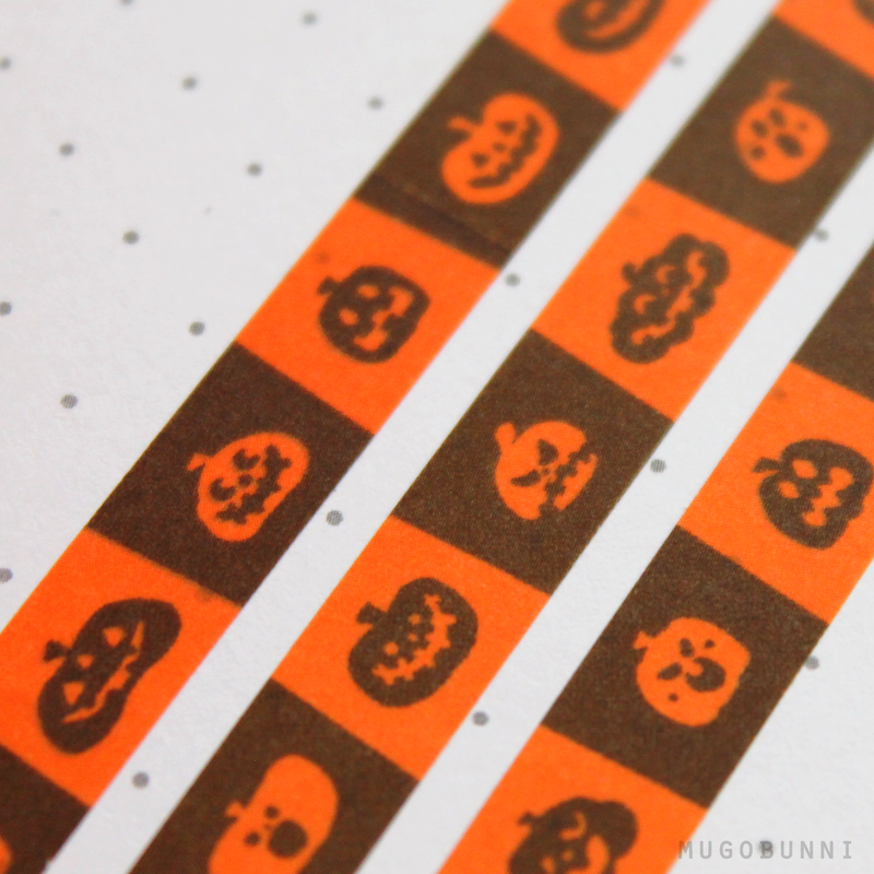 the image is a closeup shot of two pieces of halloweenthemed washi tape lying on a dotted white surface. the tape features a repeating pattern of orange and brown squares, with small black jacko'lantern silhouettes printed on each square. the overall style is cute and slightly spooky, perfect for fall crafting or decorating.  the image is welllit, highlighting the detail and texture of the tape. the watermark "mugobunni" is visible in the bottom right corner.