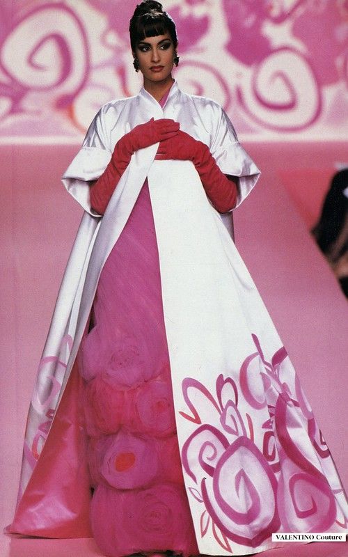 a model is walking the runway in a valentino couture fashion show. she is wearing a long, pink dress with a white overcoat that has a floral design on it. she is also wearing pink gloves. the background is a pink stage with a floral pattern.