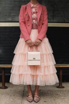 a woman in a pink blazer and a matching tulle skirt.  she is holding a small, white handbag and wearing pink shoes.  she is smiling softly and looking at the camera.  the image is lit from the front and the background is out of focus.