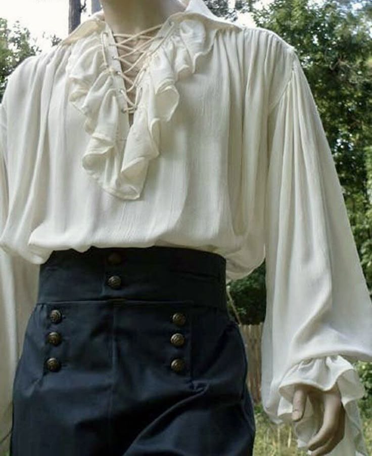 a torso shot of a person dressed in vintagestyle clothing. they are wearing a creamcolored, longsleeved shirt with ruffles around the collar and a laceup neckline. the shirt is paired with highwaisted, dark blue pants with multiple rows of buttons. the style of the outfit evokes historical periods such as the renaissance or a pirate era. the background shows hints of greenery.