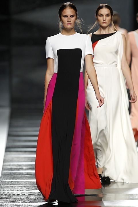 two models are walking down a runway. the model in the front is wearing a white top, black panel, and pink and red panels for her skirt. the model behind her is wearing a white dress with an offtheshoulder neckline. they are both posed in walking positions and both look elegant with their dresses.