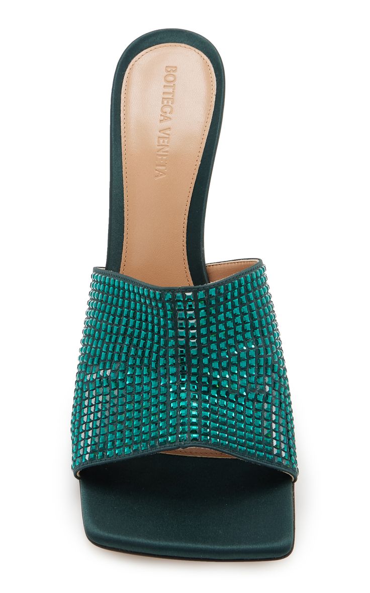 a pair of emerald green bottega veneta satin mules with a square toe and crystal embellishments are shown in this image. the shoes are presented against a white background.  the insole shows the bottega veneta logo.