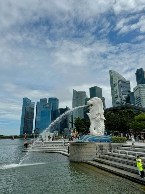 the image shows the merlion statue in singapore, with a fountain spraying water in front of it.  the background features a striking skyline of modern skyscrapers under a partly cloudy sky. the water is calm, reflecting the buildings and sky. there are people visible in the park around the merlion, and a person in a safety vest can be seen to the right of the statue near the steps.  the overall scene is bright and sunny, showcasing a typical singaporean vista.