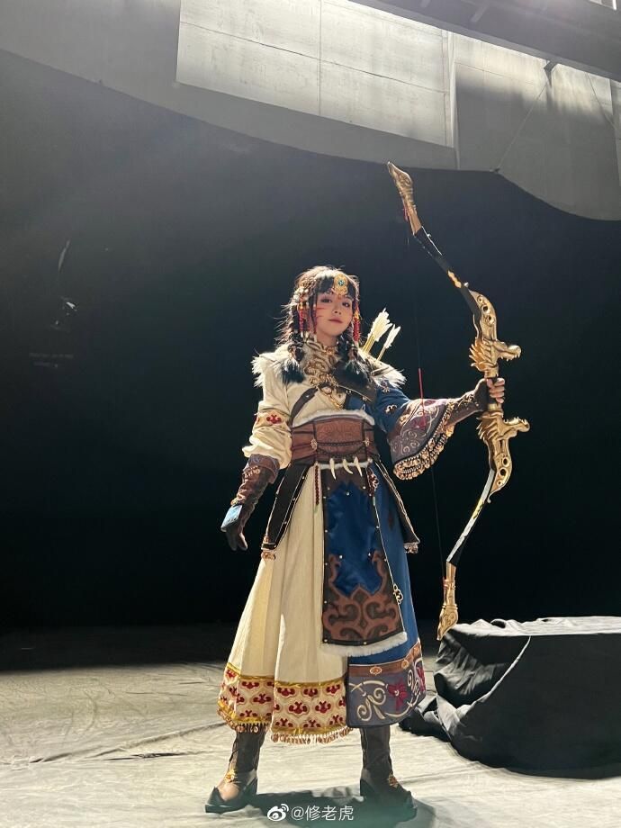 a woman in a fantasy cosplay outfit with a bow and arrow. she is posing in a studio and the background is black. the lighting is bright and there are shadows. she is wearing a long white dress with gold and red embroidery, brown pants, and a blue and gold sash. she has a gold belt, a gold necklace, and gold jewelry. she is also wearing makeup and has her hair up.