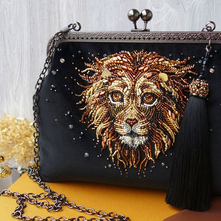a black beaded purse with a lion head design is laying on a yellow surface. the purse is made of velvet and has a gold chain. the lion's head is beaded with gold, silver, and black beads. there is a black tassel hanging from the side of the purse.