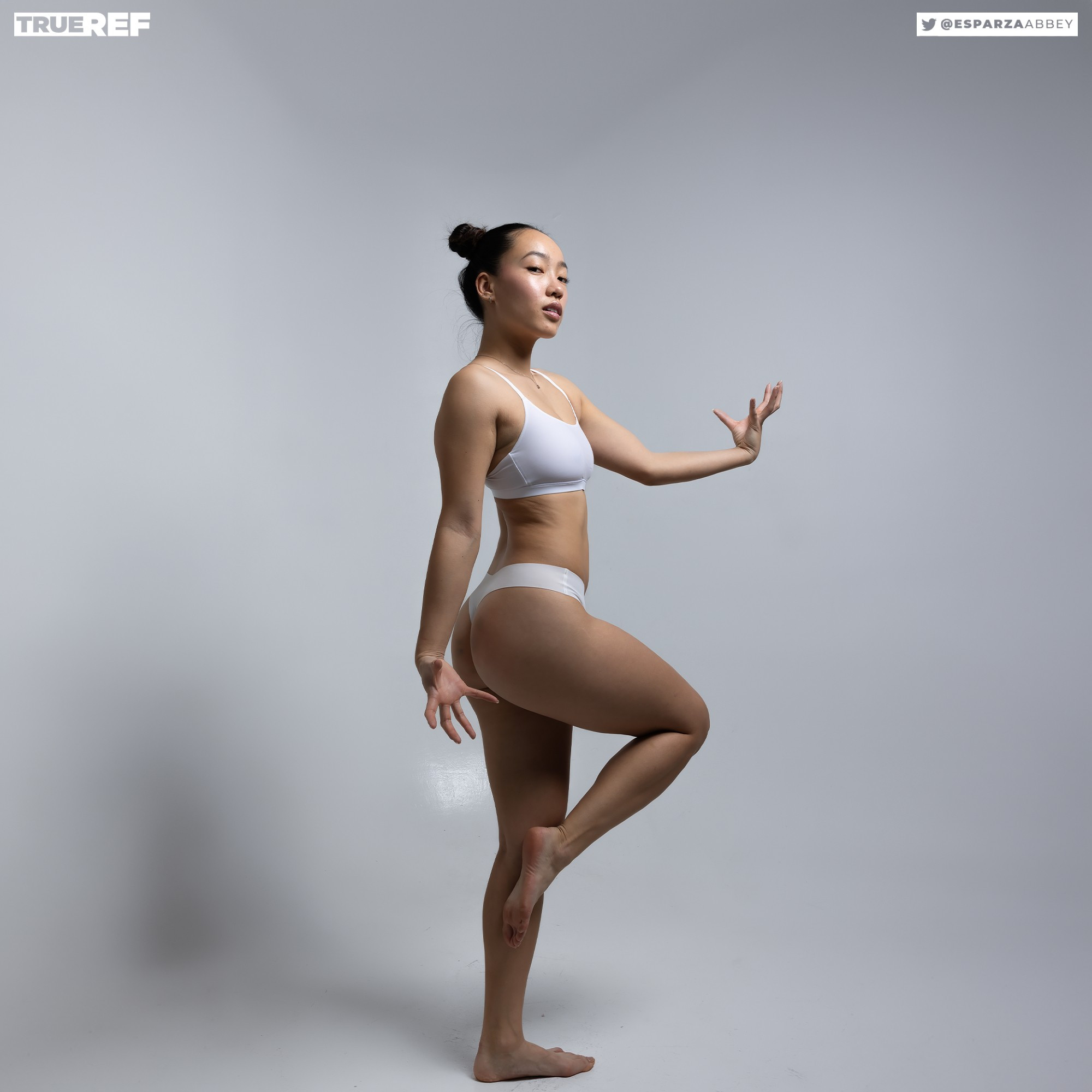 a fulllength side profile shot of a young, slender asian woman in white underwear, posing gracefully against a light grey studio background. her hair is tied up in a bun, and she has a confident expression on her face, looking slightly away from the camera. the overall aesthetic is minimalist and elegant, highlighting the model's physique and posture.