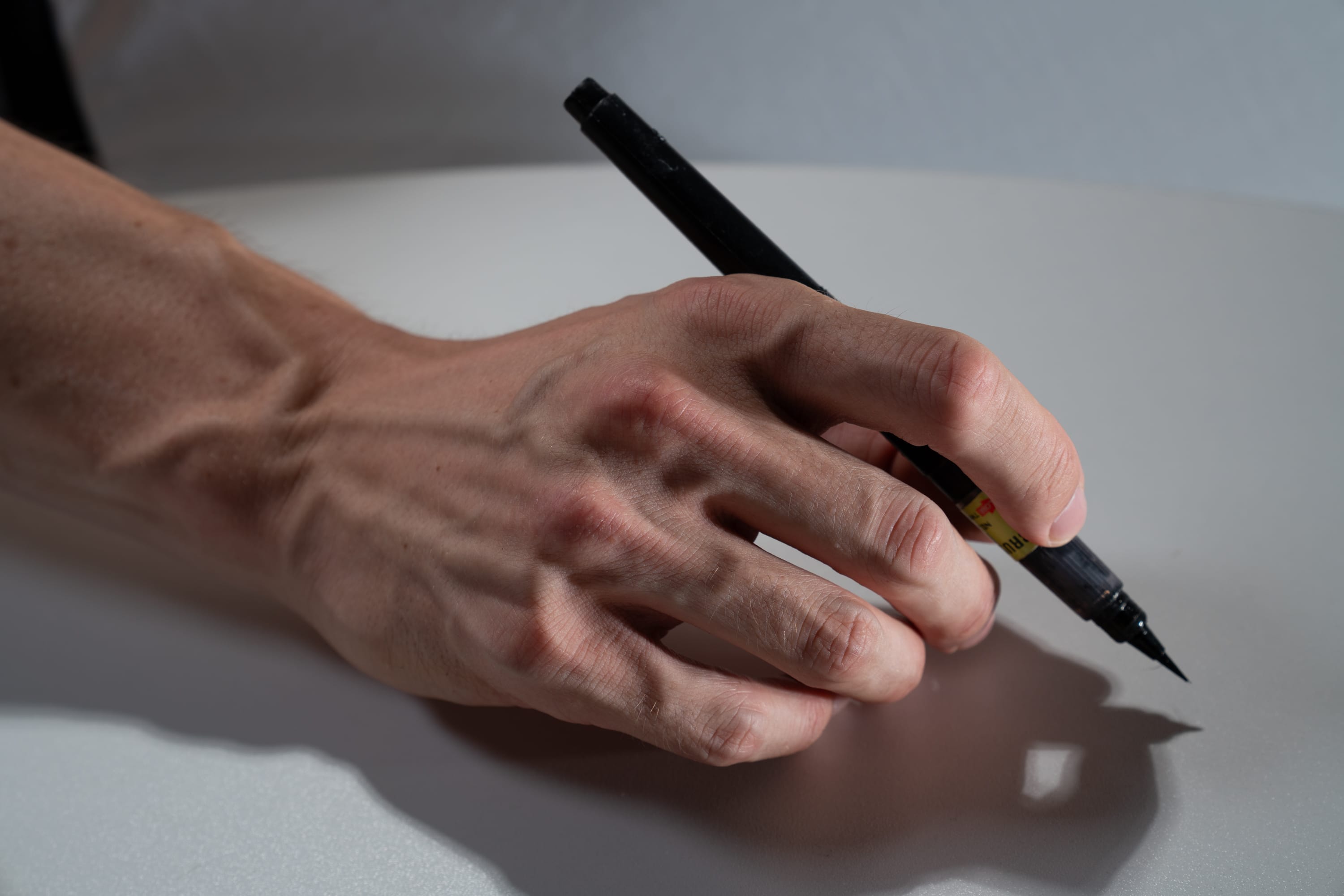 a closeup of a fairskinned hand holding a black brush pen poised above a white surface as if about to write. the hand is illuminated by studio lighting, creating distinct highlights and shadows, emphasizing the details of the skin, veins, and the pen itself.