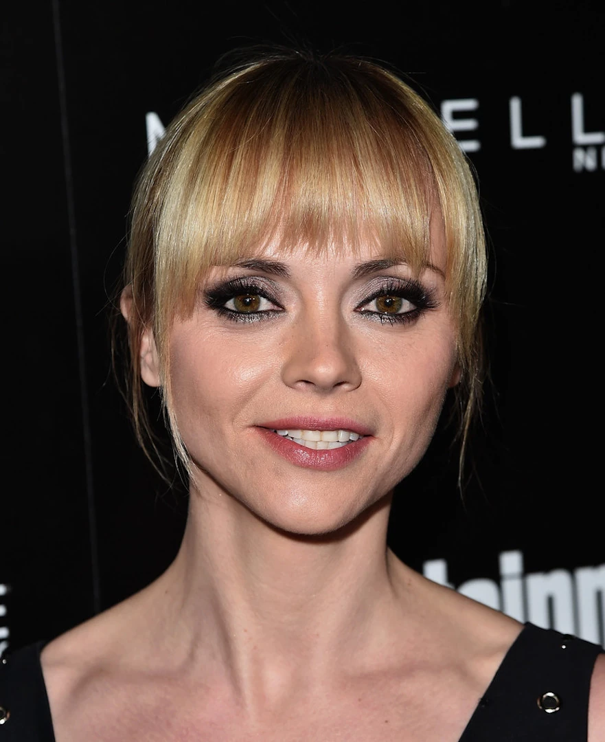 a closeup portrait of actress aubrey plaza. she is facing the camera with her hair styled in a blunt cut with bangs that fall to her eyebrows. she has light blonde hair, dark eyes with subtle eye makeup, and is wearing a black dress. the background is dark with blurred text suggesting a red carpet event or awards show. the focus is entirely on aubrey plaza's face and upper body. the image conveys a sense of sophistication and elegance.