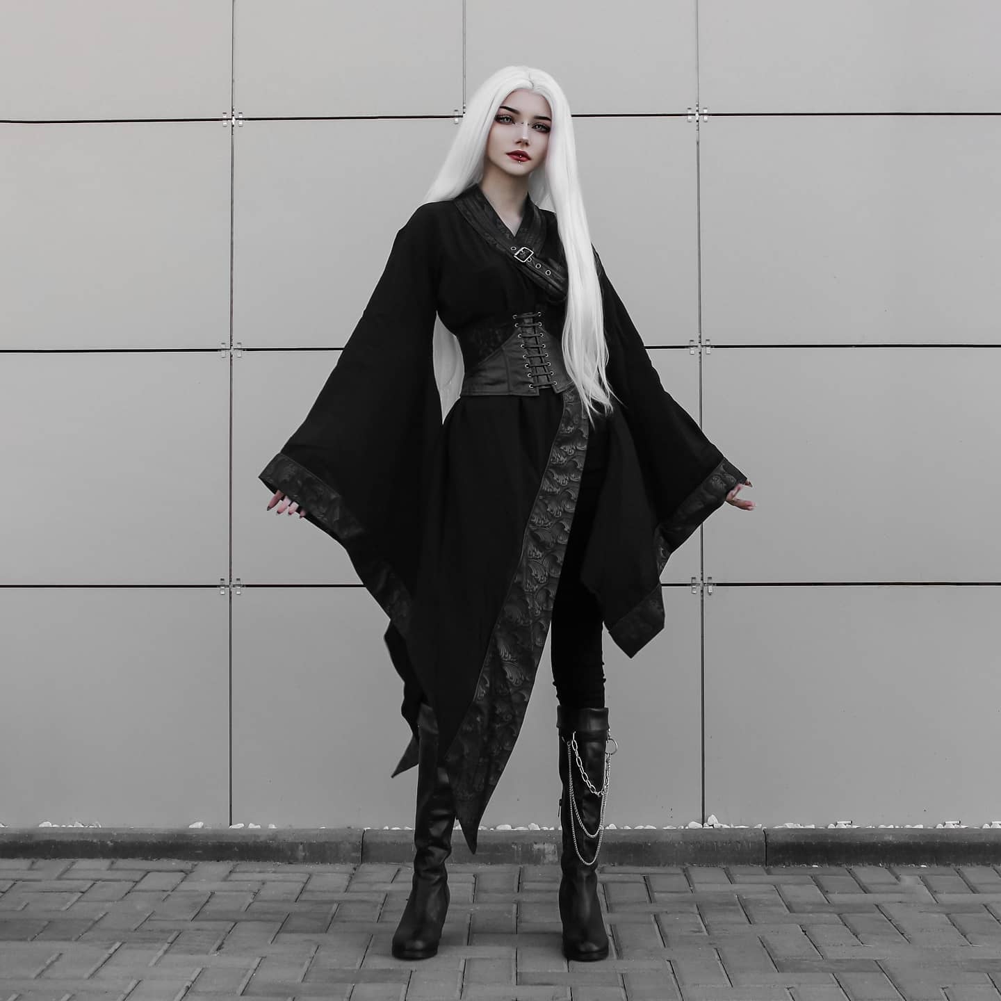 a full shot of a pale woman with long white hair modeling a black gothic kimonostyle coat, black corset, and black boots. the photo is taken outdoors against a grey wall.  the overall aesthetic is dark, edgy, and fashionforward.
