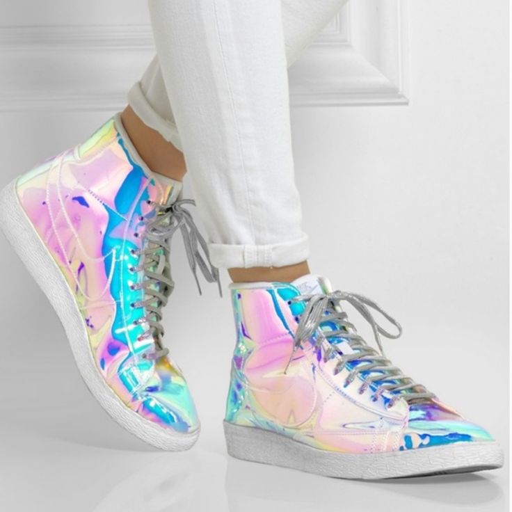 a woman is wearing a pair of iridescent nike high top sneakers. the shoes are silver with a holographic finish that makes them appear to change color in the light. the woman is wearing white pants and is standing on a white surface.