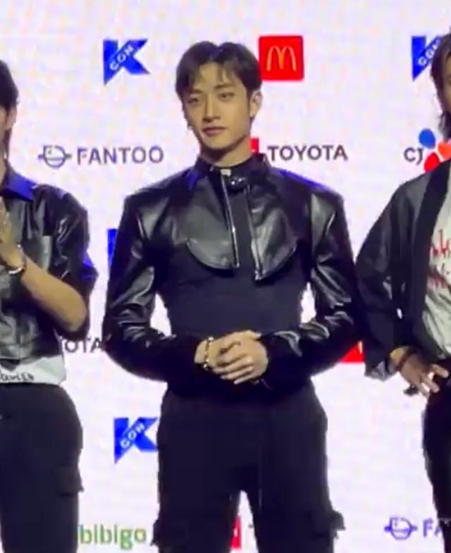 the image shows three young men, likely members of a kpop band, at what appears to be a press conference or red carpet event.  the central figure is prominently featured, wearing a stylish black cropped leather jacket over a dark top and pants.  he has short, dark hair. the men on either side of him are also dressed in dark clothing.  various sponsor logos are visible in the background including fantoo, toyota, and mcdonald's, indicating a significant event. the overall mood is formal yet fashionable.
