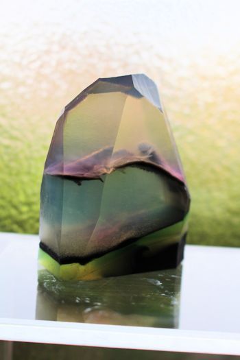 a closeup image of a handcrafted soap bar shaped like a crystal. the soap is clear and translucent, with layers of green, blue, purple, and black. it is sitting on a white surface and reflecting the light. the soap is surrounded by a white background, which enhances the colors of the soap. the soap is a beautiful and unique piece of art.