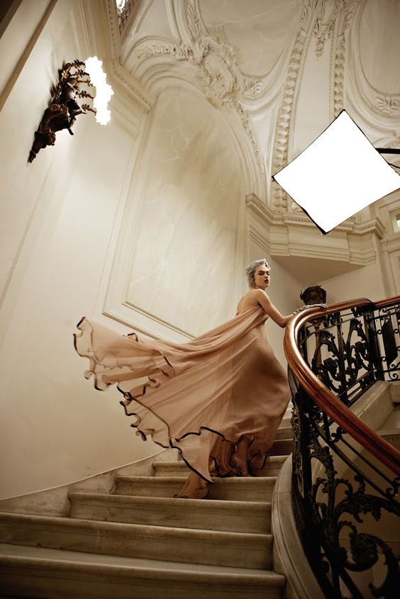 a woman in a flowing peach dress walks down a grand staircase. she is posing for a photoshoot. the image is lit with both natural light and professional lighting. the perspective is from a low angle, looking up at the woman. the overall aesthetic of the image is luxurious and elegant.