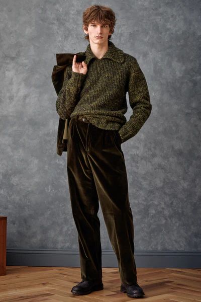 a full shot of a male model showcasing autumn/winter menswear fashion. he's wearing a green speckled knit sweater, dark olive corduroy trousers, and dark brown leather boots. he's holding a dark green jacket over his shoulder. the background is a textured dark grey wall and herringbone wood floor. the overall aesthetic is sophisticated and relaxed.
