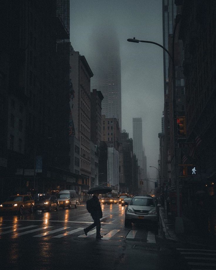 the image depicts a rainy night scene on a city street, possibly in new york city, given the architecture.  the atmosphere is dark, moody, and atmospheric, with a thick fog or mist hanging in the air.  the street is wet, reflecting the lights of passing cars. a lone man with an umbrella is crossing the street in the midground. tall skyscrapers loom on either side of the street, contributing to the overall feeling of urban density and isolation.  the lighting is low, emphasizing the contrast between the light and dark areas of the scene. the overall color palette is muted and dark, contributing to the somber mood.