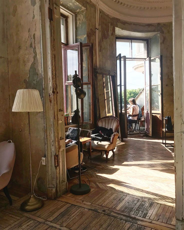 the image shows the sunlit interior of a vintage cafe or similar establishment located in an old building with rustic charm.  the room features worn, wooden parquet floors, large windows that let in abundant natural light, and peeling paint on the walls, creating a shabbychic ambiance.  old, distressed furniture, including armchairs and a floor lamp, is scattered throughout the space. two people are visible sitting near a window, one seemingly reading or working on a laptop. the style is bohemian and retro, hinting at a unique, possibly european, setting with a city view visible through the windows. the overall feeling is one of cozy comfort and relaxed atmosphere.