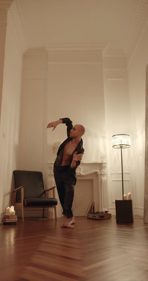 a man in a black jacket and trousers is dancing in a room with hardwood floors. he is bald and barefoot. the room is lit by a floor lamp and candles, and there is a fireplace and an armchair in the background. the image is artistic and moody, capturing the grace and expression of the dancer.