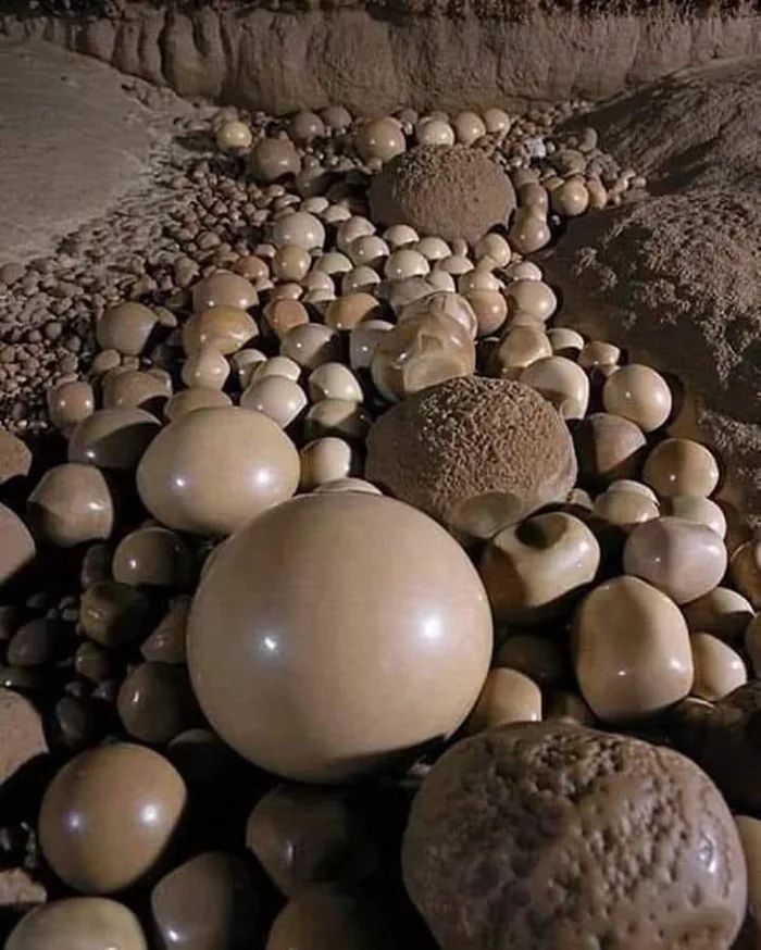 a close up image of many cave formations,  looking like large round marbles or stones of various sizes.  the formations are  shades of brown, beige and white.  these formations are the result of  mineral deposits over many years.  a large, smooth spherical formation  in the center dominates the frame.  the background is a blur of many more  formations and rock walls.