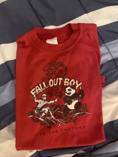 a red tshirt with the fall out boy logo on it. the logo features a panda with a sword and a ninja. the shirt has the words "fall out boy" and "live the fantasy" on it. the shirt is a vintage style, likely from the 2000s or 2010s.