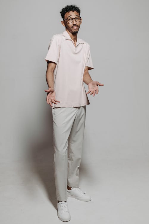 a full shot of a man standing in a studio against a white background. he is wearing glasses, a pink polo shirt, light gray pants, and white sneakers. he has a confused expression on his face and his hands are out to his sides.