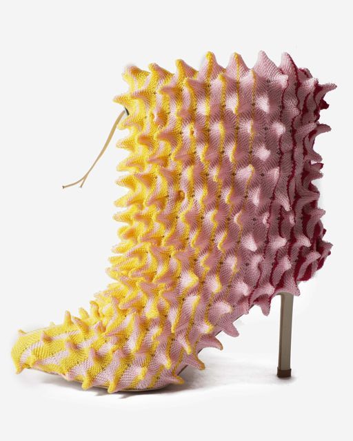 a closeup shot of a unique and unusual highheeled boot. the boot has a textile covering with a spiky texture in shades of yellow, pink, and red. the color gradually transitions from yellow at the toe to pink in the middle and red near the heel. a small, beige string protrudes from the side of the boot. the heel is a simple, silvercolored cylinder. the background is a plain white.