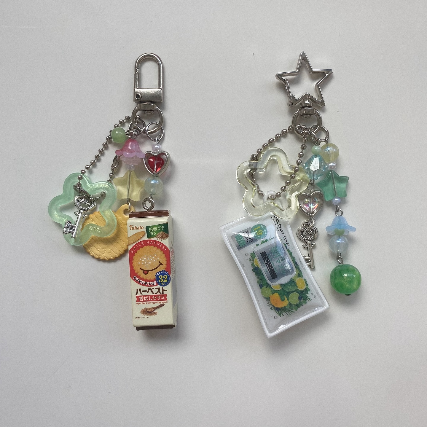 two keychains with charms on a white surface. the left keychain has a silver dshaped clasp and charms including a flower, disc, heart, key, and a miniature tobato harvest biscuit pack. the right keychain has a silver starshaped clasp with charms including a flower, heart, key, and a transparent plastic pack with an image of citrus fruits. both keychains have various beads in shades of green, blue, and white.