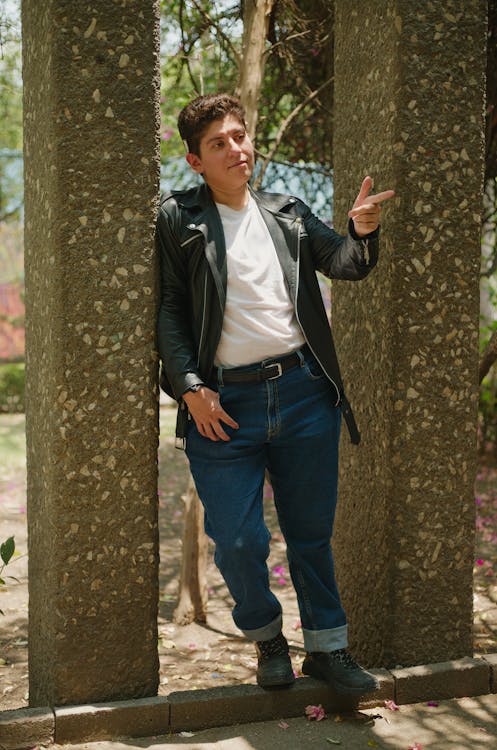 a young man stands leaning against two large stone columns in a park setting. he's wearing a black leather jacket, white shirt, blue jeans, and darkcolored boots. he's smiling and gesturing with his right hand. the sun is shining, and the background is blurry but shows green trees and foliage. the overall impression is one of casual, relaxed style and confident demeanor.