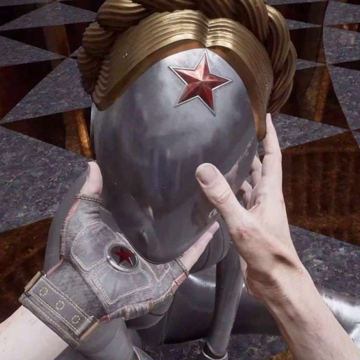 the image shows a closeup of a pair of hands gently holding a sculpted bust of wonder woman's head. the bust is unique, featuring a helmetlike mask with a prominent red star reminiscent of soviet or communist symbolism, replacing the traditional wonder woman eagle emblem. the statue is detailed, with a metallic gray finish and gold accents on the braided hair. the hands appear to be from a video game, given their slightly unrealistic texture and the overall setting's suggestive video game aesthetic. the background is a dark, polished floor with a geometric pattern, which adds to the scene's mysterious and intriguing ambiance.  the overall impression is one of reverence or careful examination of a significant object.