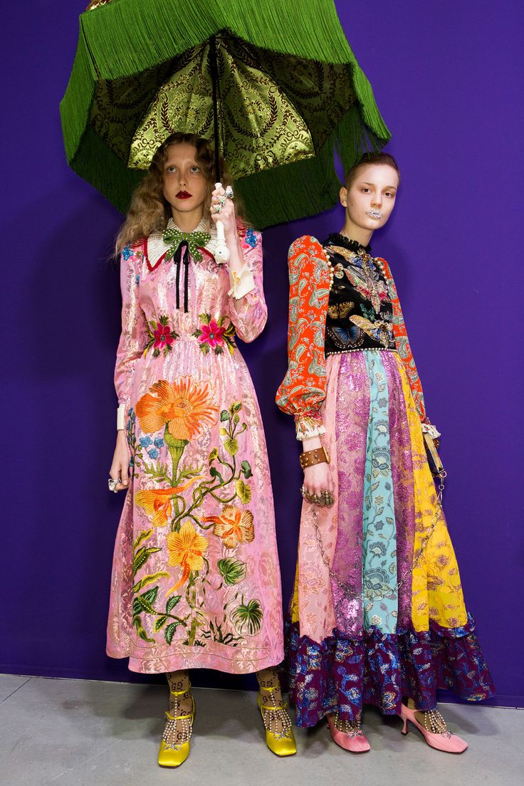 two models are showcasing extravagant gucci dresses on a purple background. the model on the left wears a pink floral embroidered dress and holds a large green fringed umbrella. the model on the right wears a multicolored patchwork dress with various patterns and textures. both models wear embellished shoes. the overall style is eclectic, blending vintage and bohemian elements with a touch of high fashion. the image is professional, likely from a fashion show or campaign.