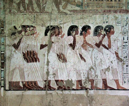 this image shows an ancient egyptian mural depicting a procession of women. the women are dressed in long white dresses and are carrying offerings. the background is a pale yellowbrown color and the outlines of the figures are black. the overall palette is muted and earthy, reflecting the pigments available in ancient egypt. this type of scene was common in tomb decoration, representing the deceased's journey to the afterlife and the abundance they would experience there.