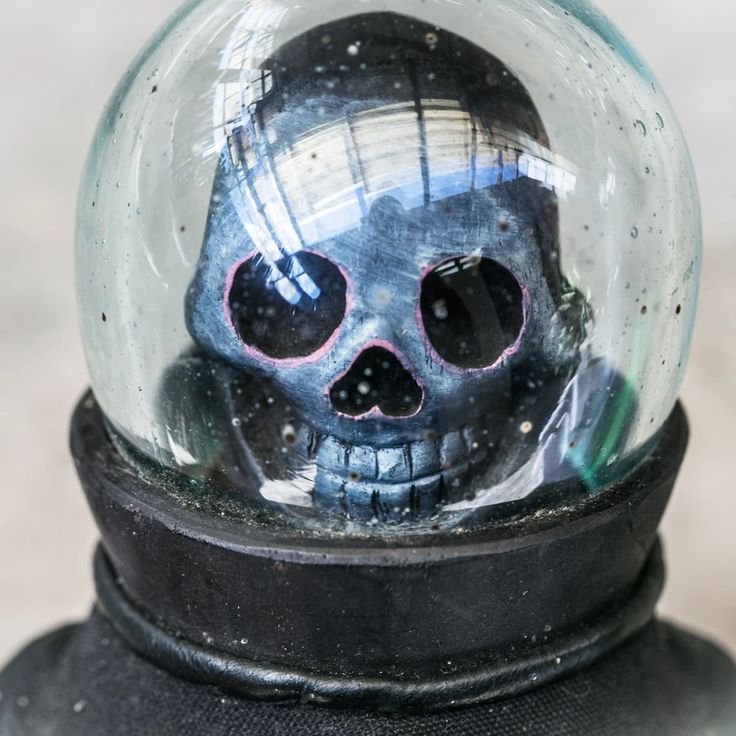 a closeup of a glass dome ornament shaped as a skull. the skull is wearing a spacesuit and has a black helmet with a clear glass dome. the skull is painted in black and white with silver accents. the background is a blurred white.